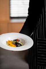 A chef wearing a striped apron serves a gourmet dish of two-toned black and yellow stuffed pasta with a creamy sauce and red caviar topping in a fine dining restaurant setting.