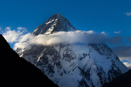 View of K2 mountain's snow-capped peak piercing the blue sky, partially veiled by soft, white clouds, contrasting with the dark foreground, Skardu, Gilgit, Pakistan.