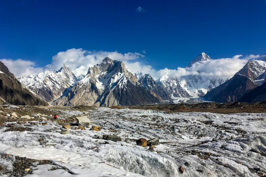 View of jagged, snow-dusted peaks piercing the azure sky above a glistening, icy expanse, a tiny red figure braving the glacial terrain, Skardu, Gilgit, Pakistan.