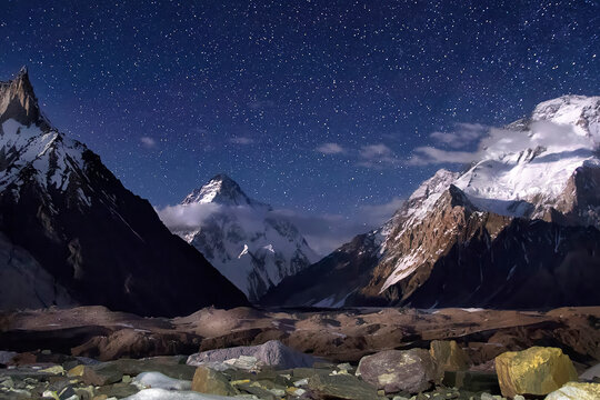 View of rugged, dark mountains and a starry sky over a rocky ground, dusted with snow, with bright white peaks piercing the night, Skardu, Gilgit, Pakistan.
