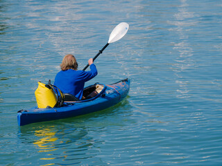 Fototapeta premium Kayaker in Blue Kayak Paddling Across Calm Blue Water With Yellow Gear. Sit-on-top kayak paddles across tranquil blue water, carrying a yellow dry bag. Ideal for outdoor sport, adventure, fitness, and