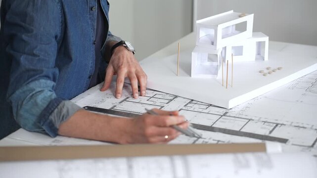 Architect sketching building plans on paper using a ruler and pen while working digitally on a laptop with a blueprint software displaying a floor plan