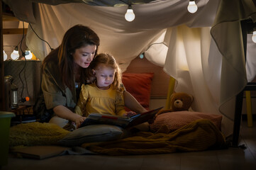 Mother reading a bedtime story to her young daughter inside a cozy blanket fort at home, glowing...