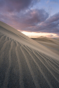 View of undulating sand dunes under a dramatic, cloudy sky, textured by wind and bathed in the soft light of the setting sun, Maspalomas, Canary Islands, Spain.