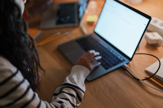 A person with dark hair, wearing a striped sweater, is typing on a laptop at a wooden desk.