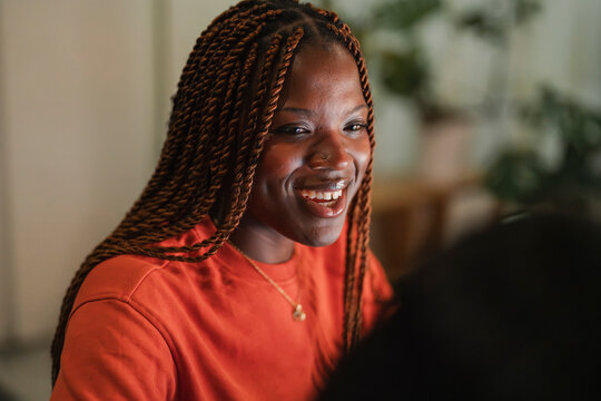 A young woman with braided hair smiles warmly while looking off to the side. She wears an orange sweatshirt and a delicate necklace.