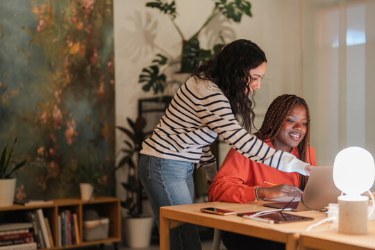Two women collaborate on a project at a desk, one pointing at a laptop screen while the other takes notes.