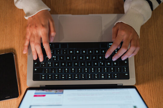 A person's hands type on a laptop keyboard, with a smartphone and tablet nearby on a wooden desk.