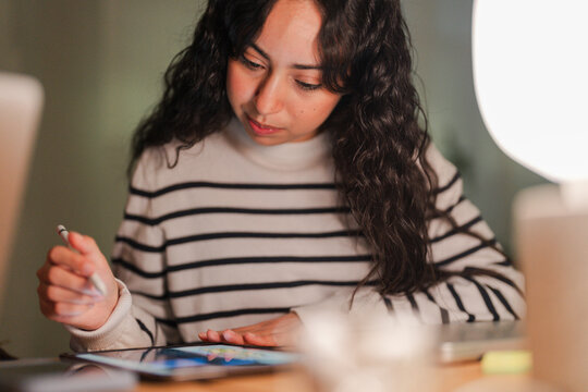 A young woman with dark, curly hair concentrates on her tablet, holding a stylus in her hand as she works or studies at a desk.