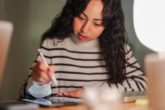 A young woman with dark curly hair concentrates on her tablet, using a stylus to draw or write.