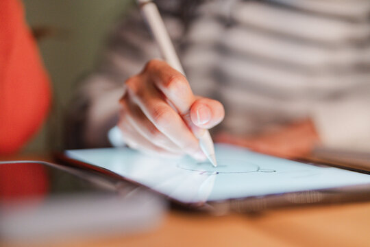 A person's hand uses a stylus to draw on a digital tablet, creating art with focused precision in a dimly lit environment.