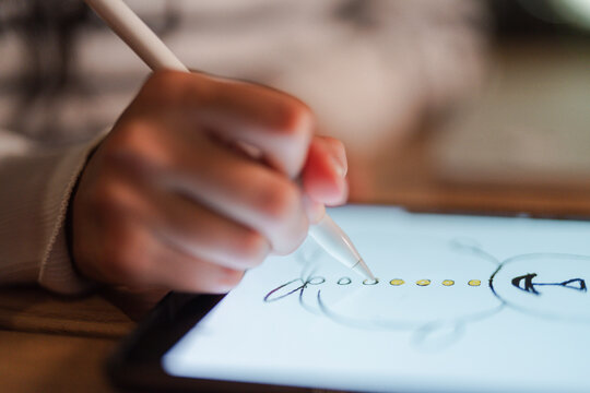 A child's hand uses a stylus to draw a snowman on a digital tablet, adding yellow dots for buttons.
