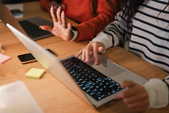 Two people collaborate on a project, one typing on a laptop while the other gestures, surrounded by sticky notes and a smartphone on a wooden desk.