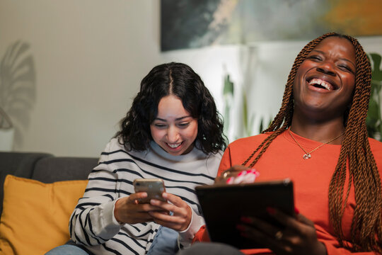 Two young women share a moment of laughter while looking at a mobile device and a tablet in a cozy living room setting.