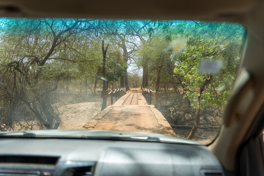 View of a weathered wooden bridge, framed by verdant trees, stretches ahead, as seen through the windshield of a vehicle, Thies, Thies Region, Senegal.