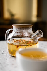 Warm-toned close-up of an amber herbal tea infusion in a modern glass teapot with a wooden lid, resting on a textured table next to a white teacup