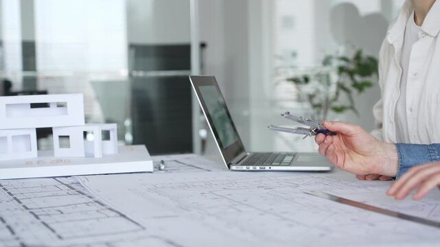 Architects collaborating over blueprints and a white scale model, using ruler and caliper on a shared desk as they plan residential and urban building projects