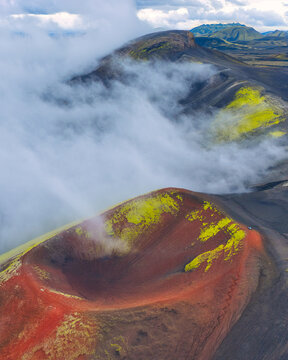 Aerial view of a volcanic landscape where red and black earth meet patches of vibrant green, partially veiled by ethereal white clouds, Akureyri, Rangarthing ytra, Iceland.