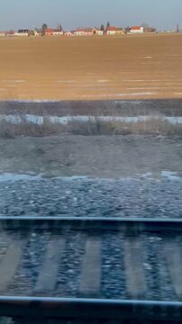train window view of tracks, ballast and sleepers with expansive brown fields beyond, rural signage and telegraph poles, motion impression with muddy verge Video taken on mobile device.