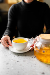 A woman in a black top holds a white cup of vibrant orange sea buckthorn (or hippophae) tea next to a glass teapot on a modern grey table