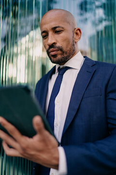 Businessman staring at tablet with calm focus, standing near glass wall, embodying modern concentration and technology-driven decision-making in corporate workflow.