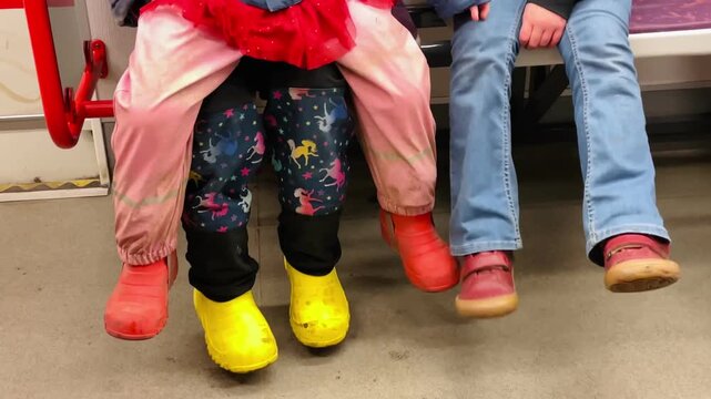 child yellow boots seated between adults on subway seat, red coat and pink pants, patterned leggings, jeans and casual shoes nearby, protective lap and close Video taken on mobile device.