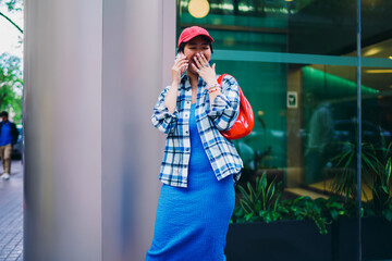 Fototapeta premium Young Asian woman laughing during phone call outdoors, wearing red cap, plaid shirt, blue dress and red bag, standing next to modern glass building, joyful street moment.