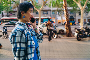 Asian woman laughing while talking on smartphone outdoors, enjoying lively conversation during a walk in the city, expressing joy, freedom, and digital connectivity in daily life. © BullRun