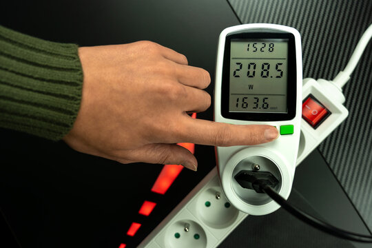 hand pointing at wattmeter, device that helps monitor electricity consumption (in watts). Amount of electricity used and the approximate cost. The red light reinforce the message