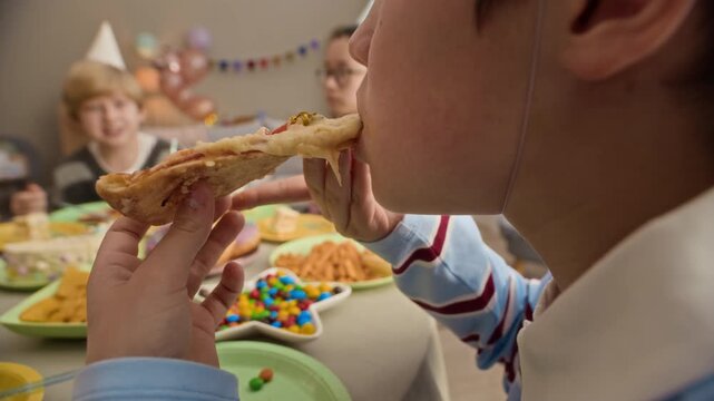 Close up of preteen Caucasian boy sitting at festive table served with fast food and biting slice of pizza with gooey chees