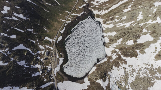 Aerial view of a partially frozen lake mirroring the sky's pale hues, bordered by rocky terrain dusted with snow, a silent sentinel in the alpine heights, Colle del Nivolet, Valle d'Aosta, Italy.