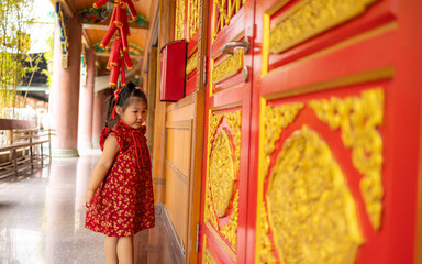 Obraz premium Adorable Little Girl in Traditional Red Cheongsam Standing by Golden Carved Temple Doors,A cute young Asian girl wearing a festive red Chinese dress (Qipao) with gold floral patterns,