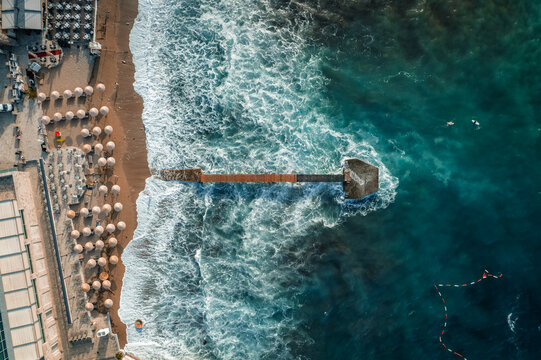 Aerial view of the textured beach, with its neat rows of umbrellas meeting the turquoise, frothy sea near the pier, Budva, Budva Municipality, Montenegro.