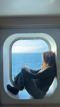 Vertical shot of a young woman sitting inside a porthole of a yacht or cruise ship while calmly observing the sea flowing outside. 