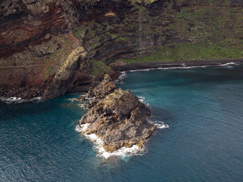Aerial view of waves crashing around a rocky outcrop against the backdrop of layered cliffs meeting a dark sandy beach Playa de Nogales, La Palma, Canary Islands, Spain.