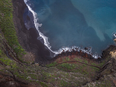 Aerial view of a dramatic coastline where dark volcanic sands meet the turquoise ocean, fringed by a narrow path clinging to the rugged cliffs, Playa de Nogales, La Palma, Canary Islands, Spain.