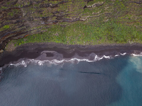 Aerial view of the rugged coastline where dark volcanic sands meet the turquoise ocean beneath a cliff adorned with vibrant green vegetation, Playa de Nogales, La Palma, Canary Islands, Spain.
