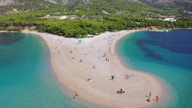 Aerial view of Zlatni Rat beach on Brac Island, Croatia. Famous pebble sand spit stretching into turquoise Adriatic Sea with tourists, pine forest and mountains in background.