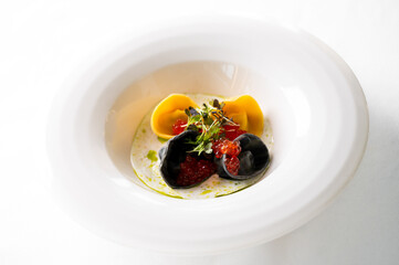 A gourmet dish of delicate yellow and black pasta (likely ravioli or tortelli) with caviar, herbs, and a creamy white sauce, presented on a fine white plate in a studio setting.