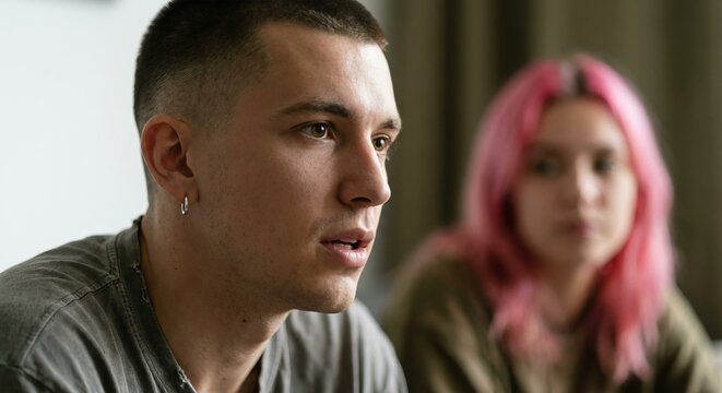 Young man with a buzz cut and earring has a serious, contemplative expression during a difficult conversation with a woman blurred in the background