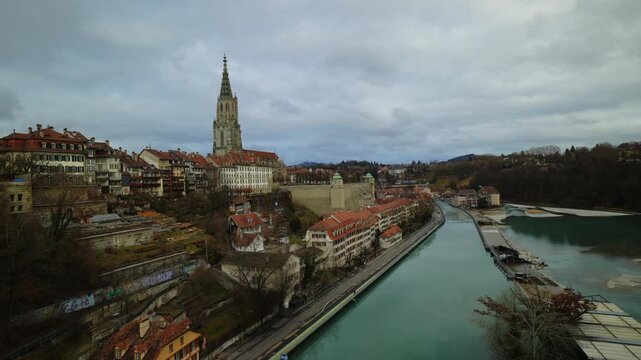 The towering gothic spire of the Bern Minster rises above the historic riverfront houses and the emerald waters of the Aare, captured under a dramatic canopy of gray clouds that emphasize the timeless