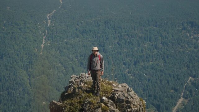 Steadying hiker shifting on narrow peak, scanning valley with red backpack and wide-brim hat