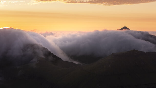 Aerial view of a sea of clouds rolling over rugged mountains, a solitary peak piercing through the misty veil under a golden sky, Torshamn, Faroe Islands.