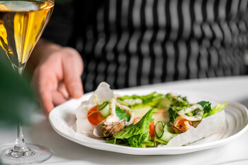 A close-up shot of a gourmet seafood salad with shrimp, clams, fresh greens, cucumber, and tomatoes, served in rice paper on a white plate held by a chef in an apron. A glass of wine is nearby