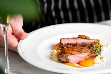 Close-up of a perfectly cooked, sliced duck breast on a white plate with orange pur&eacute;e, baby carrots, and microgreens in a fine dining restaurant setting