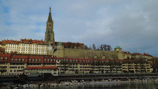 The towering gothic spire of the Bern Minster rises above the historic riverfront houses and the emerald waters of the Aare, captured under a dramatic canopy of gray clouds that emphasize the timeless