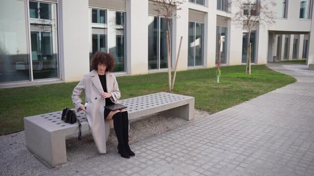 A Stylish Woman is Sitting and Waiting Comfortably on a Bench in a Modern Urban Landscape
