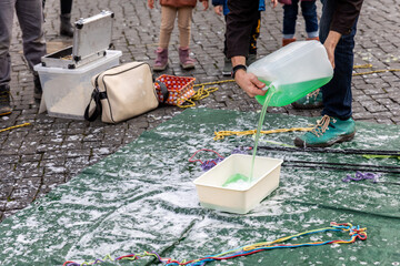 Street artist pours green bubble solution into container on pavement, tools and ropes around, showing preparation before performance. Urban scene captures manual process, creativity street art workup © Kyryl Gorlov
