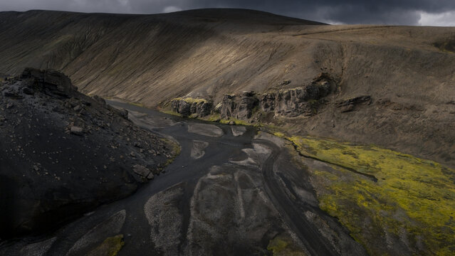 Aerial view of a river carving through a stark landscape, where dark volcanic rock meets vibrant green moss under a brooding sky, Iceland.