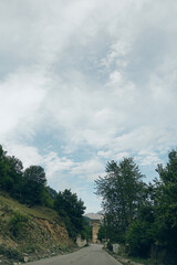 Obraz premium Road through countryside with trees and mountains under dramatic cloudy sky, scenic nature landscape and empty route for travel and exploration
